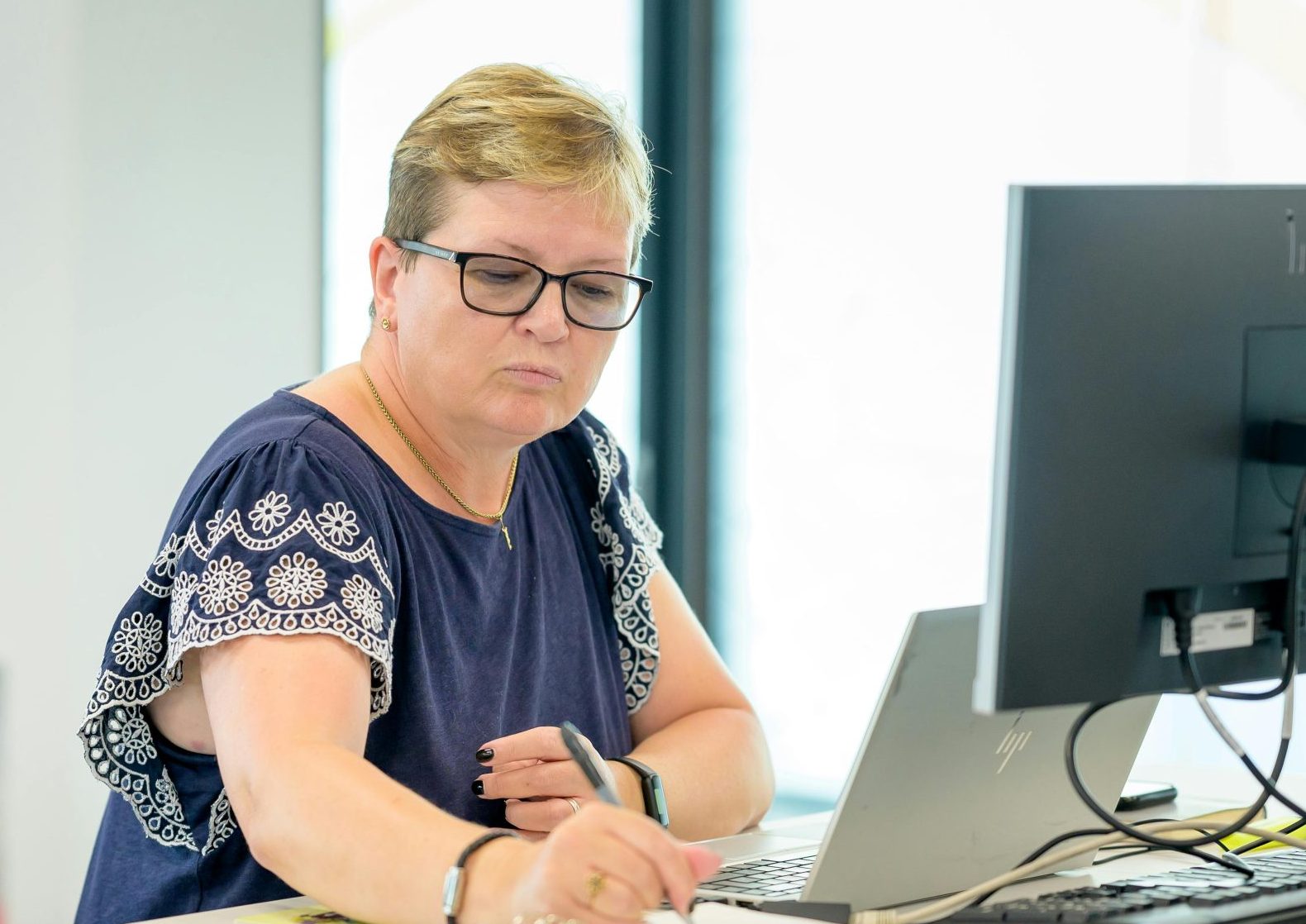 Professional woman wearing eyeglasses working at an office desk with a laptop and notebook.